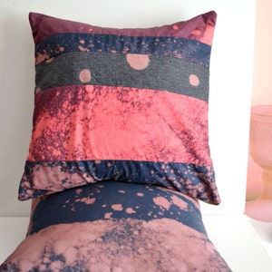 May include: Two decorative throw pillows with a patchwork design. The top pillow has a dark grey, pink, and blue striped pattern with white splatter paint. The bottom pillow has a dark blue and pink splatter paint pattern.