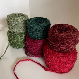 May include: A collection of yarn skeins in various shades of green and red. The yarn has a soft, textured appearance, with a fuzzy or velvety surface. The colors include olive green, forest green, burgundy, and a vibrant red. Ideal for knitting and crochet projects.