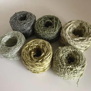 May include: Seven spools of yarn in various shades of green, gray, and gold are arranged on a white surface. The yarn appears to be a textured ribbon-like material, perfect for crafting projects. The colors range from light silver to a deep olive green.