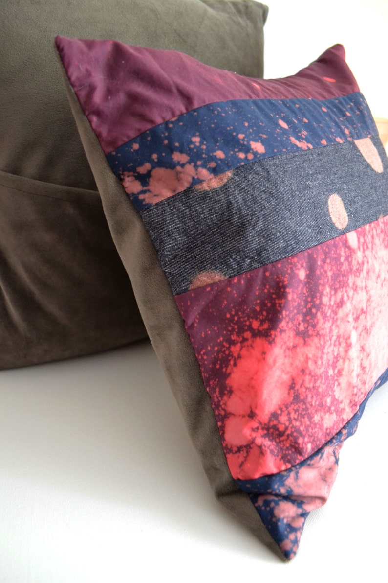 May include: A multi-colored throw pillow with a patchwork design. The pillow is made of different fabrics, including denim, velvet, and cotton. The pillow has a splatter paint design in shades of pink, blue, and white.