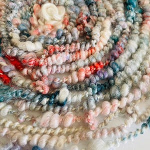 May include: Close-up of a circular arrangement of colorful yarn. The yarn features a variety of colors, including white, pink, red, blue, and gray. The yarn is thick and textured, with a slightly fuzzy appearance. The overall effect is soft and inviting.