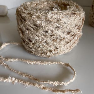 May include: A close-up of a beige yarn ball with a textured, fuzzy appearance. The yarn is cylindrical, with a strand extending from the ball. The yarn appears to be soft and suitable for knitting or crochet projects.