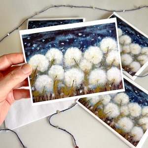 Puede incluir: Un conjunto de tarjetas de felicitación con una pintura de cabezas de semillas de diente de león blancas sobre un fondo azul oscuro con motas blancas. Las tarjetas están en blanco por dentro y son perfectas para enviar un mensaje especial.
