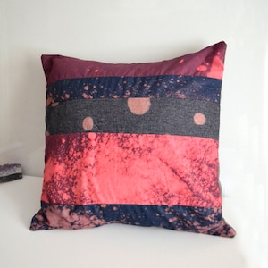 May include: A square pillow with a multi-colored striped pattern. The pillow is made of a soft, textured fabric and features a pink, blue, and gray color scheme. The pillow has a splatter paint design.