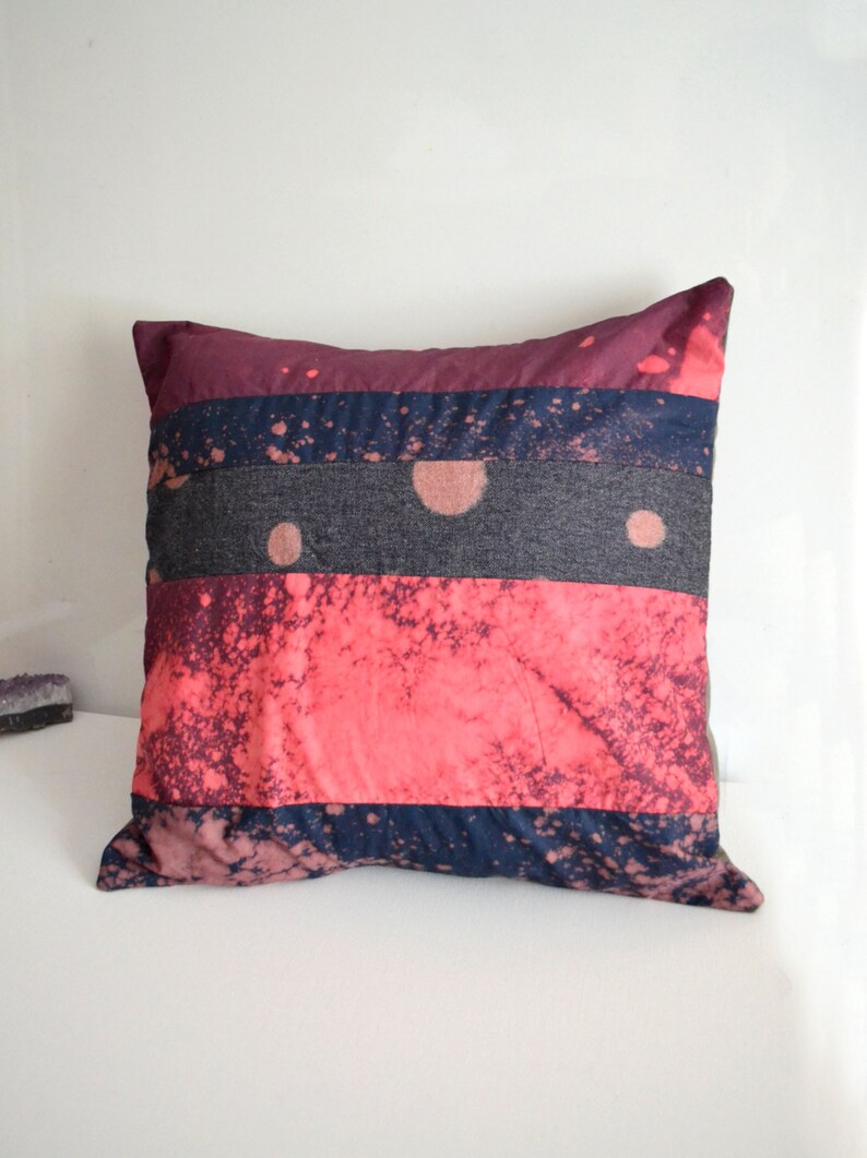 May include: A square pillow with a multi-colored striped pattern. The pillow is made of a soft, textured fabric and features a pink, blue, and gray color scheme. The pillow has a splatter paint design.