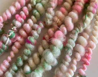 May include: A close-up of a bundle of yarn in pink, green and white colours. The yarn is soft and fluffy, and it is ideal for knitting or crocheting.