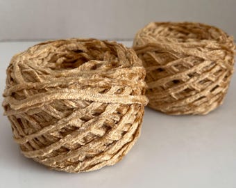 May include: Two round balls of light tan, textured yarn. Each ball is composed of a continuous strand of soft, velvet-like material wound into a dense, spherical shape with a visible crisscross pattern throughout the surface.