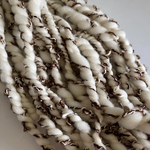 May include: A bundle of thick, cream-colored yarn twisted with a thin, dark brown textured thread. The yarn features a high-twist construction where the soft white fibers are spiraled together with the contrasting brown filament throughout.