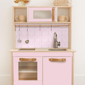 May include: A light pink wooden toy kitchen with a microwave, sink, and cabinets. The microwave and cabinet doors have tan leather-look handles. The backsplash has vertical pink and white stripes. Includes toy utensils and woven baskets.