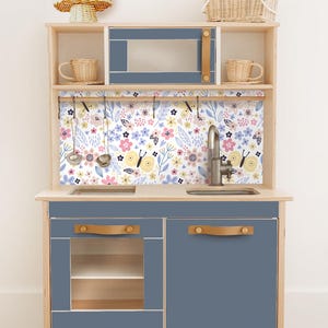 Könnte beinhalten: Eine Spielzeugküche aus Holz mit blauen und naturbelassenen Holzakzenten. Sie verfügt über eine Mikrowelle, ein Waschbecken und Schränke mit lederartigen Griffen. Die Rückwand hat ein Blumenmuster mit rosa, gelben und blauen Blumen. Inklusive Spielzeugutensilien und geflochtenen Körben.