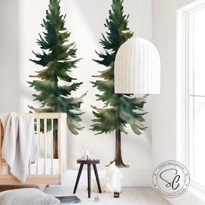 May include: A wall mural featuring two large watercolor pine trees in shades of dark and forest green with brown trunks. The mural is applied to a white wall behind a light wood crib, a small dark stool, and a white woven pendant light fixture.