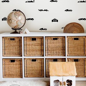 Peut inclure: Un ensemble d'autocollants muraux noirs repr&eacute;sentant diverses silhouettes de voitures de course vintage dispos&eacute;es en motif sur un mur clair au-dessus d'un meuble de rangement blanc avec huit paniers en osier et une petite maison jouet en bois.