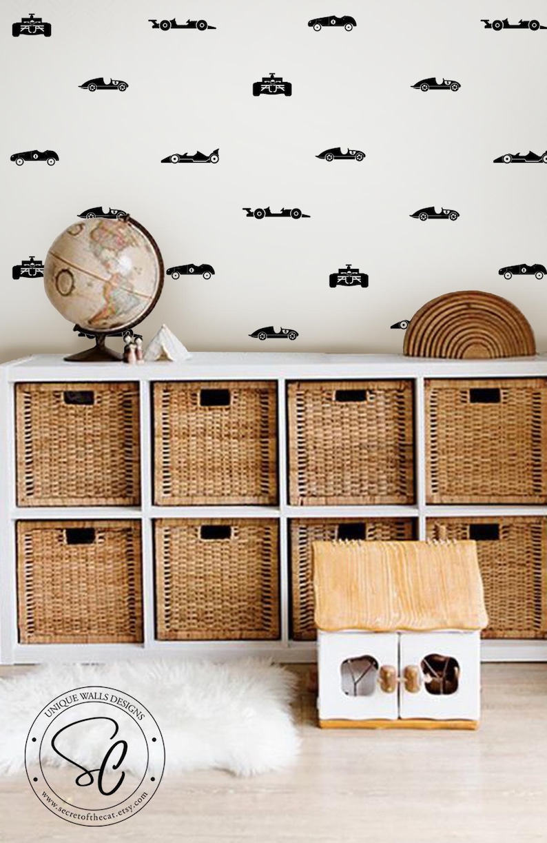 Peut inclure: Un ensemble d'autocollants muraux noirs repr&eacute;sentant diverses silhouettes de voitures de course vintage dispos&eacute;es en motif sur un mur clair au-dessus d'un meuble de rangement blanc avec huit paniers en osier et une petite maison jouet en bois.