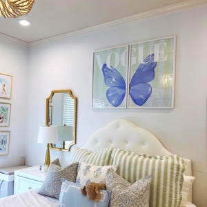 May include: Bedroom decor featuring a tufted white headboard, striped and patterned pillows, and a bedspread. Two framed prints with the word "VOGUE" and a blue butterfly design hang above the bed. A gold mirror and lamp are also visible.