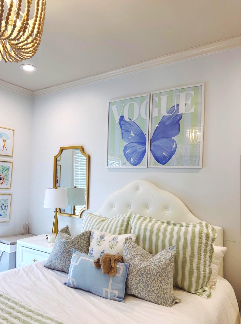 May include: Bedroom decor featuring a tufted white headboard, striped and patterned pillows, and a bedspread. Two framed prints with the word "VOGUE" and a blue butterfly design hang above the bed. A gold mirror and lamp are also visible.