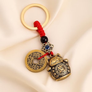 Puede incluir: Un llavero dorado con un amuleto de gato de la suerte, una moneda con caracteres chinos y una cuenta decorativa. El llavero tiene un cordón rojo y un anillo dorado.