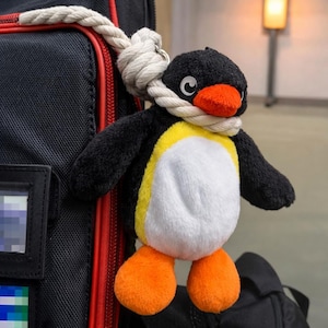 Puede incluir: Un llavero de pingüino de peluche con cabeza y alas negras, vientre blanco y detalles amarillos. Tiene pico y pies naranjas y está sujeto a una cuerda blanca. El llavero está colgado de una bolsa negra.