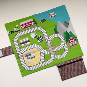 May include: A green and white play mat with a train track design. The mat features illustrations of a train station, general store, houses, trees, and a boat. A brown zippered pouch is attached to the mat.