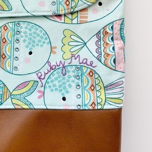 May include: A light blue fabric pouch with a brown leather bottom. The fabric features a repeating pattern of colorful fish with a whimsical design. The pouch is personalized with the name "Ruby Mae" embroidered in purple thread.