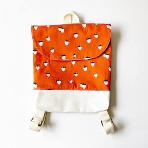 May include: A small backpack with a flap closure. The top portion is orange with a white strawberry pattern. The bottom is white. It has adjustable straps and a top handle. The backpack is designed for children.