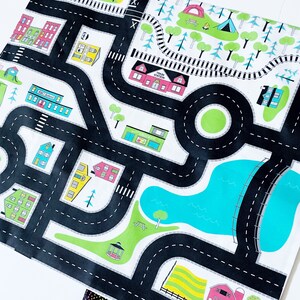 May include: A black and white play mat with a city theme. The mat features roads, buildings, a train station, a park, a lake, and trees. The mat is designed for children to play with toy cars and other vehicles.