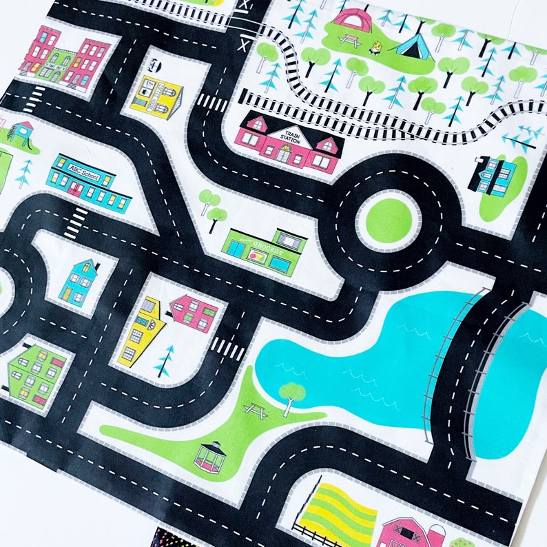 May include: A black and white play mat with a city theme. The mat features roads, buildings, a train station, a park, a lake, and trees. The mat is designed for children to play with toy cars and other vehicles.
