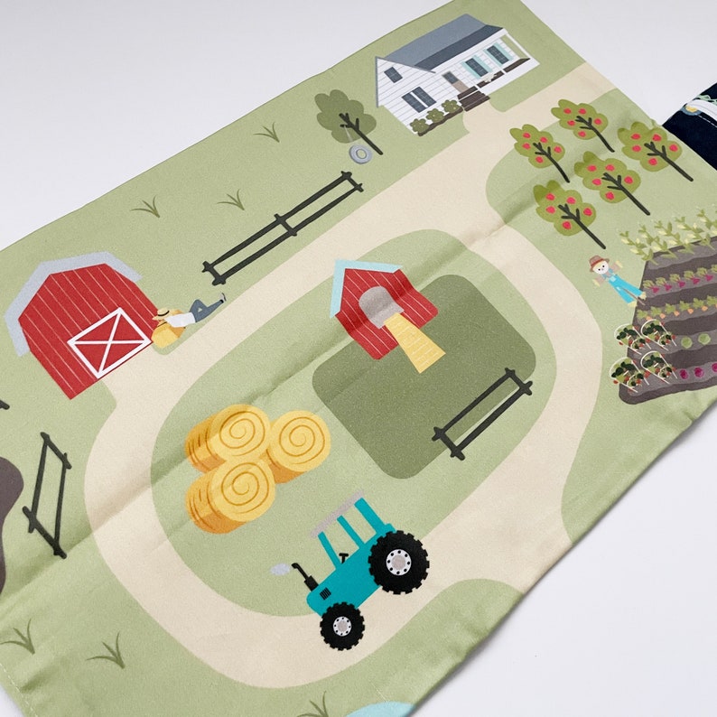 May include: A green and beige play mat with a farm theme. The mat features a red barn, a house, a tractor, hay bales, a fence, and a garden with vegetables.