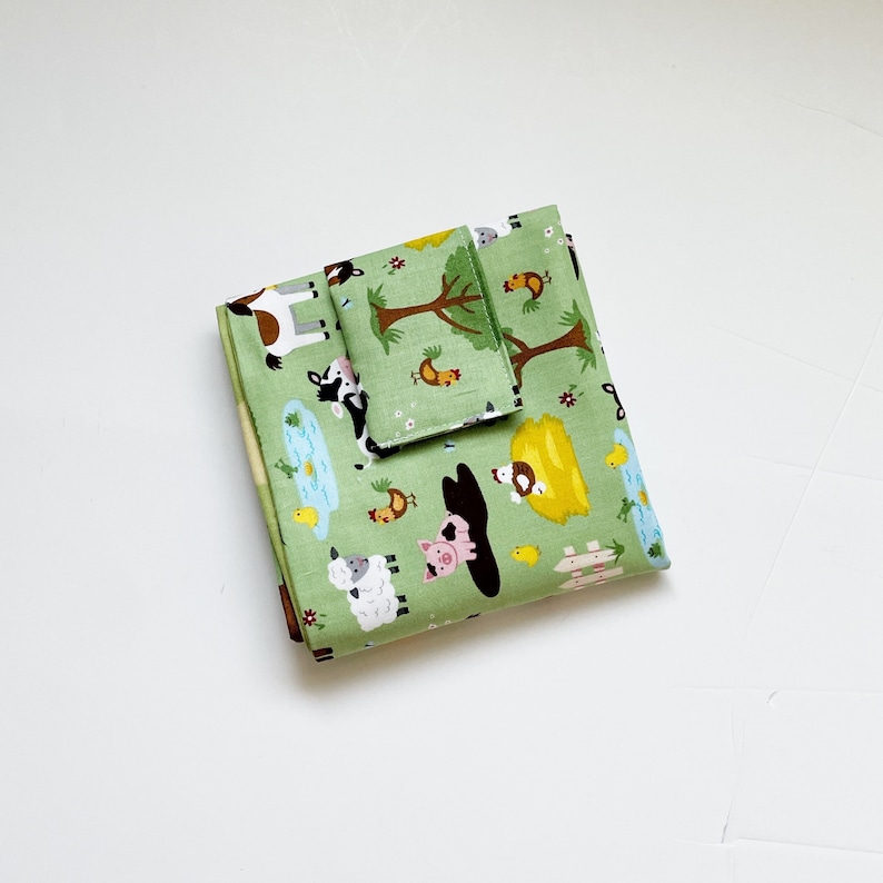 May include: Green fabric with a farm animal print. The fabric features a variety of animals including cows, pigs, chickens, sheep, and a farmhouse. The fabric is folded in half and has a white background.