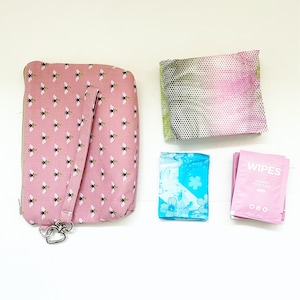 Pu&ograve; includere: Una pochette rosa con cerniera con un motivo a forma di ape e una tracolla. La pochette &egrave; accanto a un assorbente igienico con motivo floreale blu e bianco, un assorbente igienico con motivo rosa e verde e un pacchetto rosa di salviette che dice "Wipes Sensitive Pocket Size".