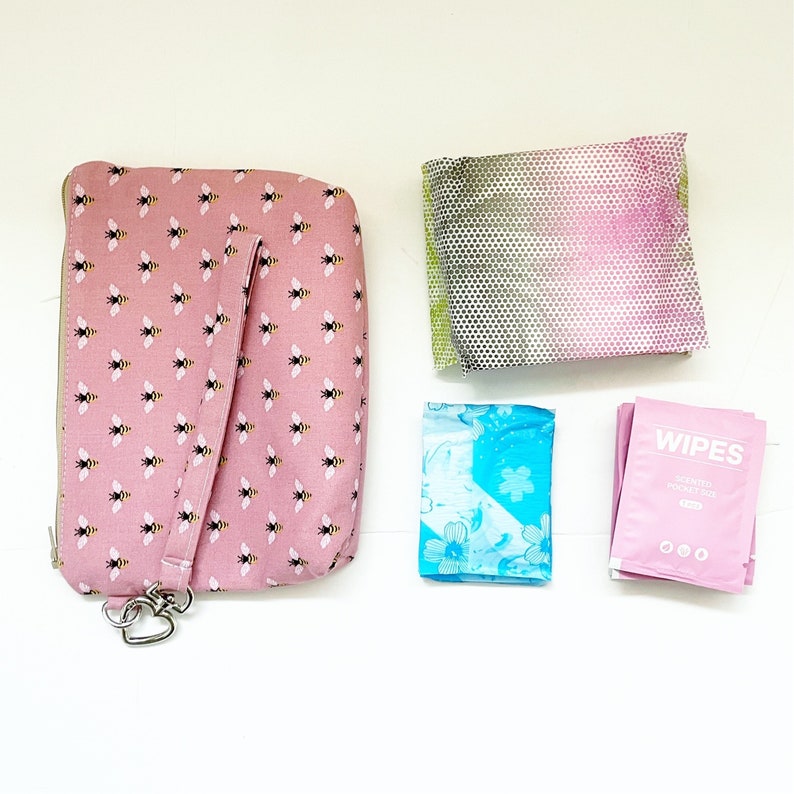 Pu&ograve; includere: Una pochette rosa con cerniera con un motivo a forma di ape e una tracolla. La pochette &egrave; accanto a un assorbente igienico con motivo floreale blu e bianco, un assorbente igienico con motivo rosa e verde e un pacchetto rosa di salviette che dice "Wipes Sensitive Pocket Size".