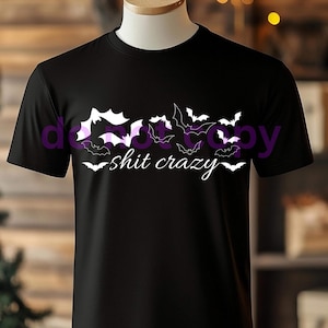 Puede incluir: Camiseta negra con un diseño de murciélagos en blanco y morado. La frase "shit crazy" está escrita en blanco debajo de los murciélagos. La camiseta se muestra en un maniquí.