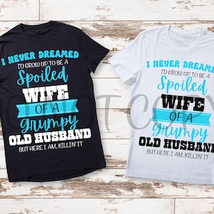 May include: Two t-shirts, one black and one white, with the same text design. The design reads "I NEVER DREAMED TO GROW UP TO BE A Spoiled WIFE OF A Grumpy OLD HUSBAND BUT HERE I AM, KILLIN' IT" in blue, white, and black.
