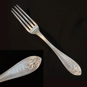 May include: A silver fork with four tines, featuring ornate floral and scrollwork detailing on the handle. The handle also has the initials "AMM" engraved. The fork is set against a dark background.
