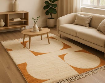 May include: A cream-colored area rug with a geometric design featuring large orange circles and semi-circles. The rug has a fringed edge and is placed in a living room setting with a sofa and coffee table.