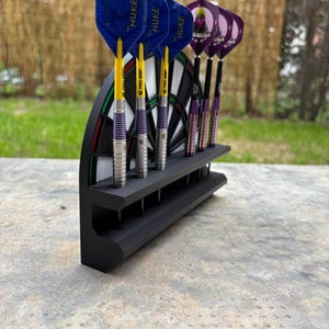 Puede incluir: Un soporte de dardos negro con una sección de diana incorporada, que contiene seis dardos. Los dardos tienen plumas azules y moradas, cañas amarillas y barriles plateados. Los dardos están etiquetados "NUKE" y "PRO GRIP".