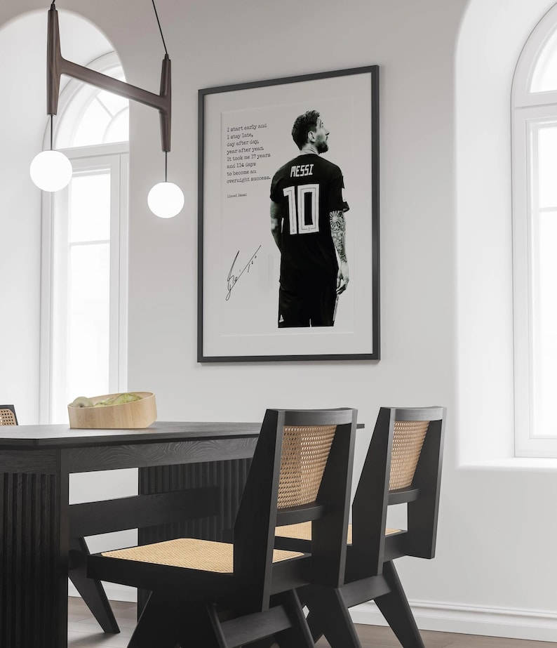 May include: Framed black and white print featuring a soccer player in a black jersey with the number 10 and the name MESSI. The print includes a quote and a signature. The print is displayed on a white wall.