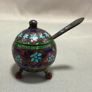 Op de afbeelding: Een kleine, ronde houder met deksel en een lange steel, voorzien van gedetailleerd cloisonné emailwerk in tinten dieprood, helderblauw en groen. Het object staat op drie korte pootjes met rode accenten en heeft een centrale knop op het deksel.