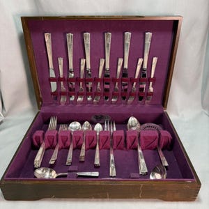 May include: A wooden flatware storage chest with a purple velvet interior lining. The set includes various silver-toned knives, forks, and spoons arranged in dedicated slots. The chest features a hinged lid and a rectangular wooden exterior finish.