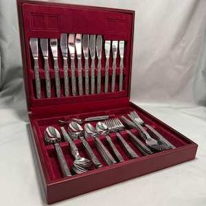 May include: A silver-toned stainless steel flatware set arranged in a dark wooden storage case with a deep red velvet interior lining. The set includes multiple knives, spoons, and forks, each featuring a decorative embossed pattern on the handles.
