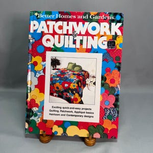 May include: A Better Homes and Gardens Patchwork & Quilting hardcover book featuring a vibrant, multi-colored floral pattern cover. The center displays a photograph of a quilt on a bed with text detailing projects, basics, and designs.