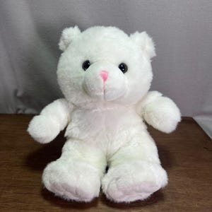 Op de afbeelding: Een pluche teddybeer met zachte witte vacht, ronde zwarte ogen en een kleine roze geborduurde neus. Het knuffeldier zit rechtop en heeft ronde oren, korte armen en dikke benen met zichtbare stiksels op de voetzolen.