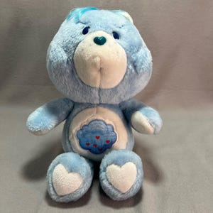 May include: A light blue plush bear with a white belly featuring a cloud design with red hearts. The bear has a white muzzle, a green heart-shaped nose, and blue eyes. The bear's feet have white heart-shaped accents.