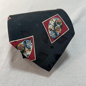 Puede incluir: Una corbata negra con un patrón de diamantes repetido. La corbata presenta dos cuadrados rojos con un diseño colorido y arremolinado en el centro. El diseño incluye acentos azules, dorados y blancos.