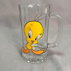 May include: Clear glass beer mug featuring a cartoon image of Tweety Bird in yellow, orange, and black. The mug has a handle and a textured base. The Tweety Bird graphic is centered on the front of the mug.