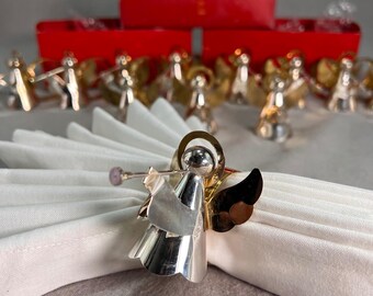 Könnte beinhalten: Eine Sammlung von Serviettenringen in Form von Engeln. Die Ringe sind aus silber- und goldfarbenem Metall gefertigt und weisen detaillierte Flügel und Heiligenscheine auf. Die Engel sind auf weißen Servietten angeordnet, mit roten Schachteln im Hintergrund. Eine festliche Tischdekoration.