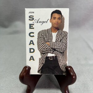 May include: A Jon Secada Angel cassette tape featuring a photograph of a person wearing a black and white checkered blazer over a white t-shirt and black pants with a black belt. The text JON SECADA and Angel is printed on the white cover.