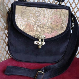 May include: Black velvet shoulder bag with a floral tapestry flap. The bag features a top handle, an adjustable strap, and a brass clasp. The tapestry displays pink, white, and gold flowers. The bag is suitable for everyday use.