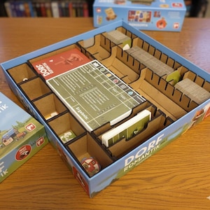 Könnte beinhalten: Eine blaue Spielbox mit einem Holzeinsatz, gefüllt mit Karten und Spielsteinen. Die Box ist mit "Dorf Romantik" beschriftet und hat ein Landschaftsdesign. Der Einsatz hat mehrere Fächer zur Organisation der Spielkomponenten.