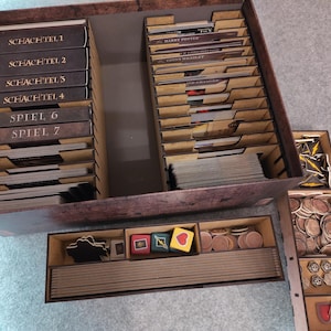 May include: A wooden board game organiser with several compartments. It contains game cards, tokens, and dice. The compartments are labelled with German text. The organiser is designed to hold a board game.
