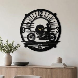 Op de afbeelding: Een ronde wandklok van zwart metaal met een silhouet van een persoon op een motorfiets tegen een zonsopgang. De wijzerplaat bevat de cijfers 3, 6, 9 en 12, met dunne goudkleurige wijzers voor de uren en minuten in het midden.